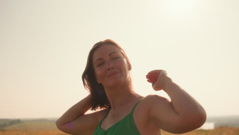 lady in green sleeveless dress stands in golden field with eyes closed and arms resting behind head, facing sunlight with peaceful expression as warm glow and summer breeze touch her skin