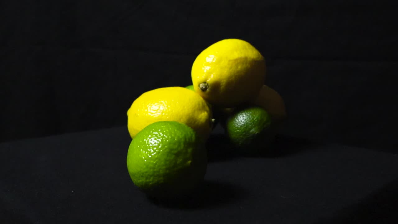 Close up footage of vibrant and colorful yellow and green limes and lemons on display on a dark black studio background while footage zooms out from them and spins around them slowly. Textured fruit.