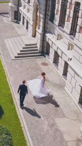 happy young bride turns around walking with groom near elegant buildings in city street on sunny day aerial view