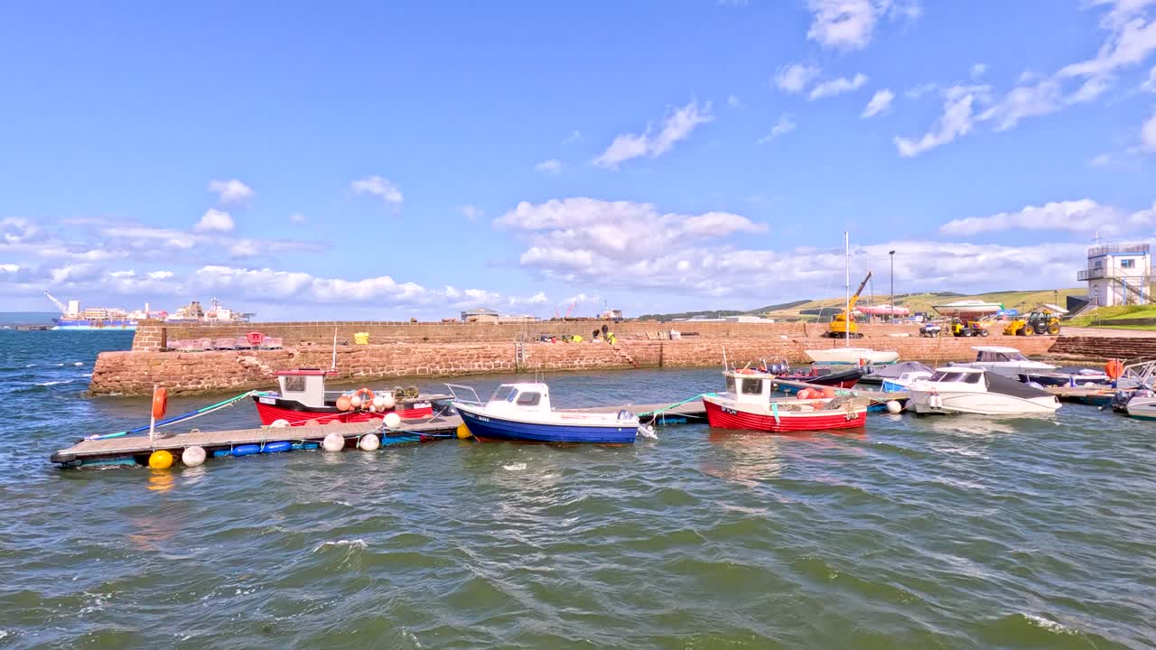 Several small boats float on choppy water beside a stone pier in Cromarty, Scotland, under bright daylight with a wide, steady camera view