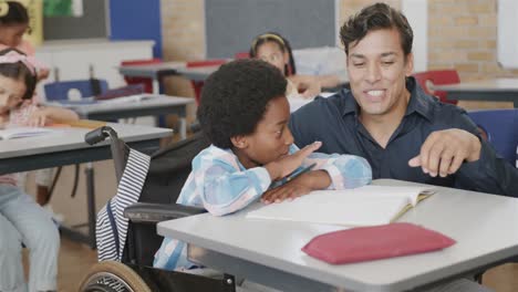 Happy-diverse-male-teacher-helping-schoolboy-in-wheelchair-reading-in-classroom-at-elementary-school