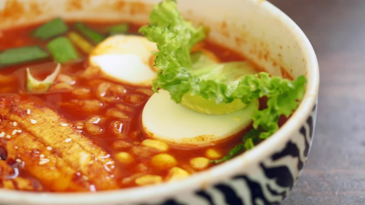 A close-up of a vibrant bowl of spicy ramen or noodle soup with eggs, green onions, and fish cake, sprinkled with sesame seeds