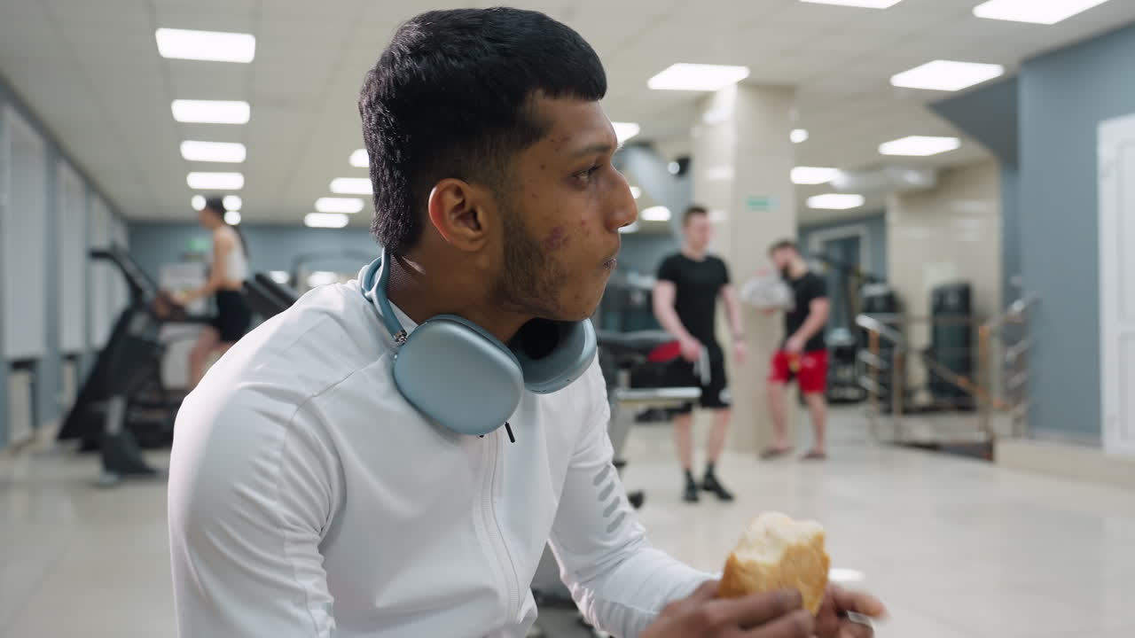 focused young man in white athletic wear with headphones around neck eats sandwich in gym setting while people in background walk, exercise, and use fitness machines in well lit indoor environment
