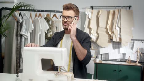 man tailor in glasses sitting at the table with a sewing machine and choosing clothes pieces and talking on the smartphone