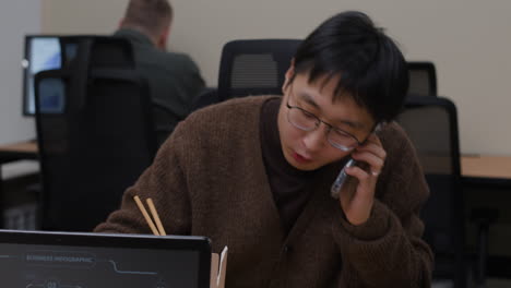 man eating with chopsticks while using cell phone in office