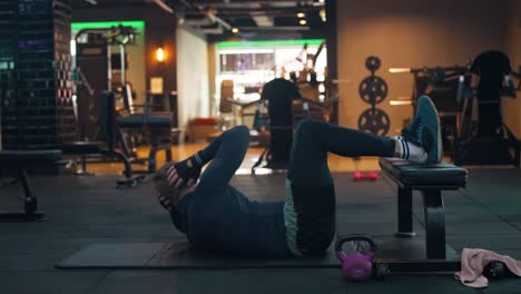 fit man performs oblique twist crunches with legs resting at 90 degrees on a bench in a dimly lit gym - hands behind head, sportsman twist his torso to reach knee with opposite elbow engaging obliques