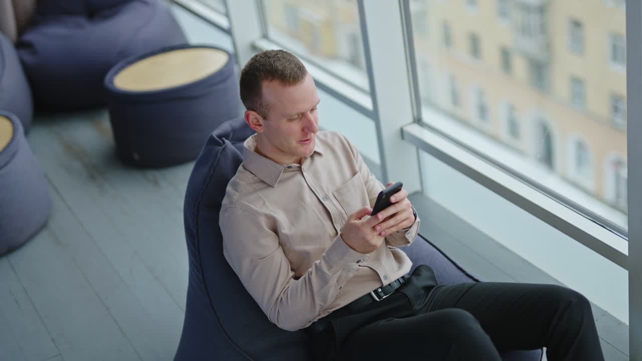 Young man holding a phone and texting to someone. Male employee sitting in comfortable armchair during lunch break. View from above.