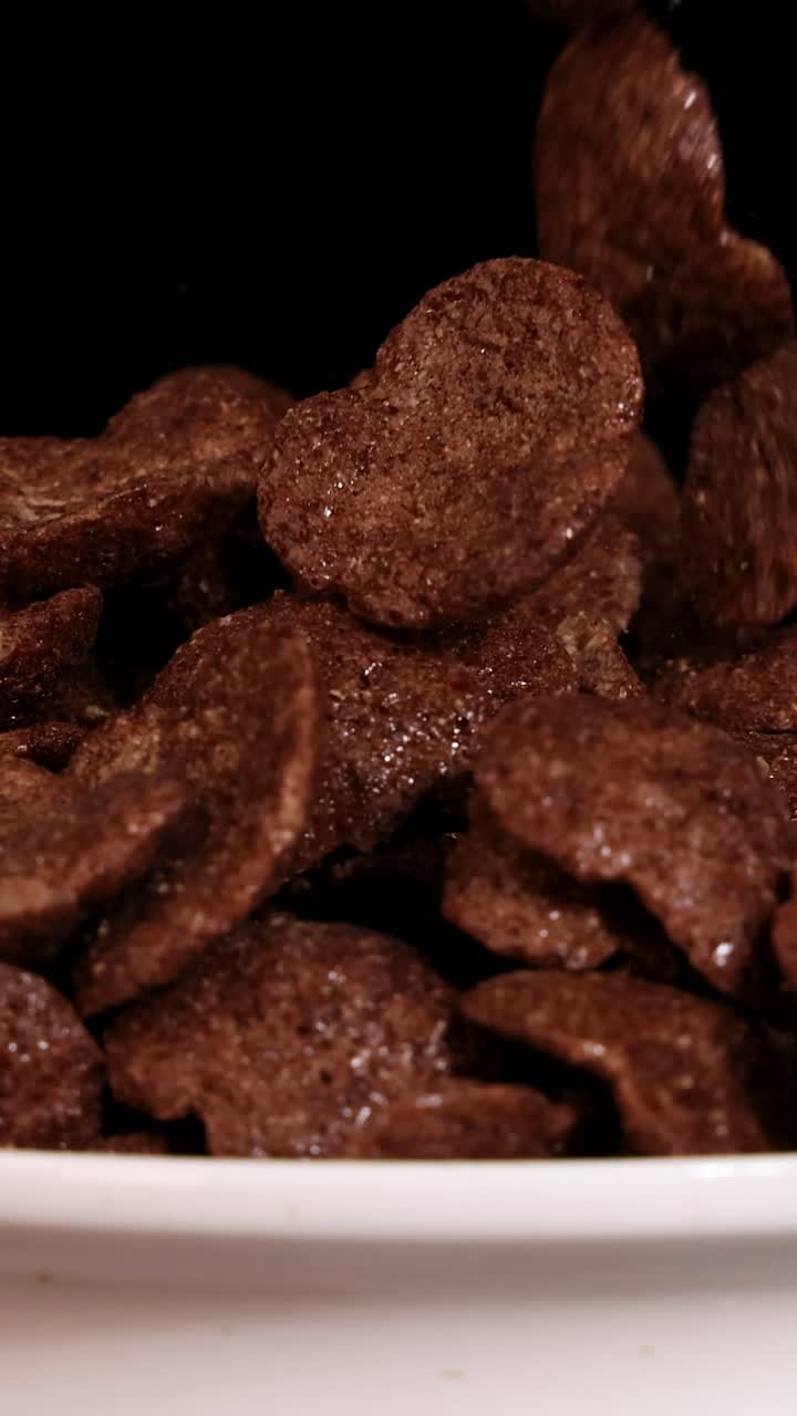 Fiocchi di cereali al cioccolato che cadono in una ciotola bianca su uno sfondo scuro, evidenziando texture e movimento in primo piano