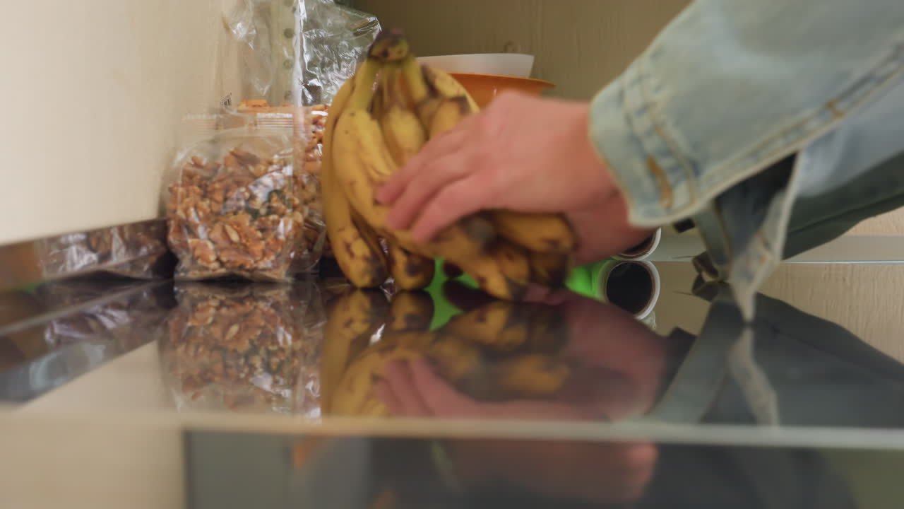 Plátanos maduros sobre una encimera de acero reflectante junto a cuencos verdes y una bolsa de cereales; una mano con manga vaquera se extiende para coger un racimo; una cálida mañana hogareña; una esquina ordenada de la despensa; reflejos sutiles y acero inoxidable.