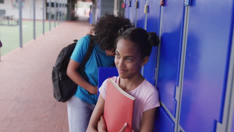 video of two diverse, happy schoolgirls talking by lockers in school corridor, copy space