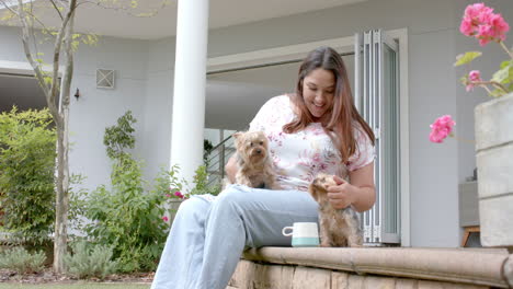 happy plus size biracial woman sitting in garden with coffee, petting dogs, copy space, slow motion