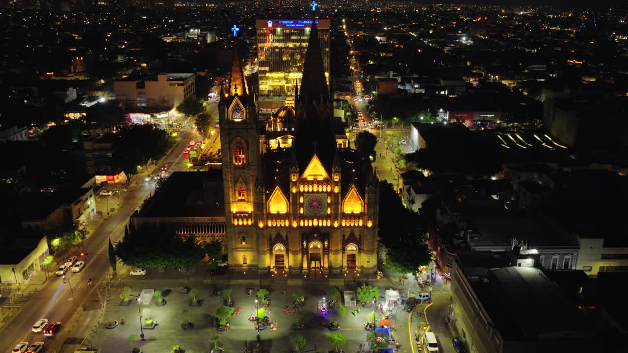 Illuminated Templo Expiatorio del Santísimo Sacramento at night with plaza, streets, and city lights in Guadalajara, Mexico. Dolly in aerial view
