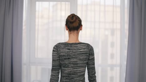 Back-view-of-young-attractive-woman-opening-curtains-on-a-big-window-and-letting-the-light-in-the-room.-Looking-out-the-window