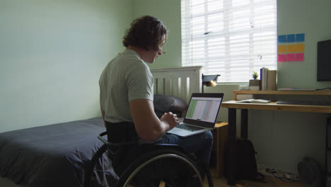 hombre con discapacidad caucásico enfocado en silla de ruedas usando computadora portátil en el dormitorio