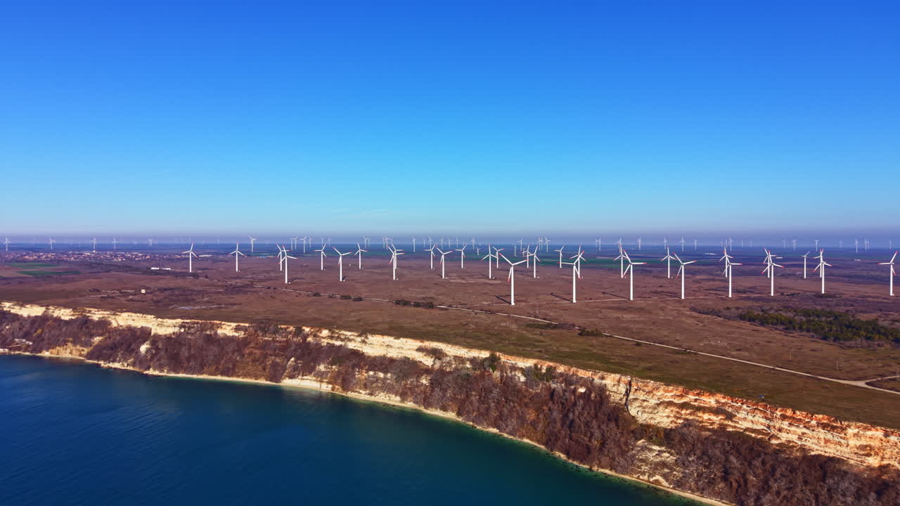 Wind turbines stand along the shoreline near a large body of water in a flat landscape on a clear day