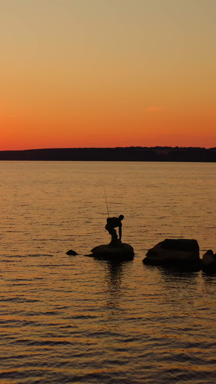 River at sunset. Silhouette of a fisherman on stone. Man going away from the lake after fishing on orange sky background in the evening. Vertical video
