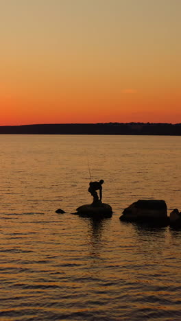 river at sunset. silhouette of a fisherman on stone. man going away from the lake after fishing on orange sky background in the evening. vertical video