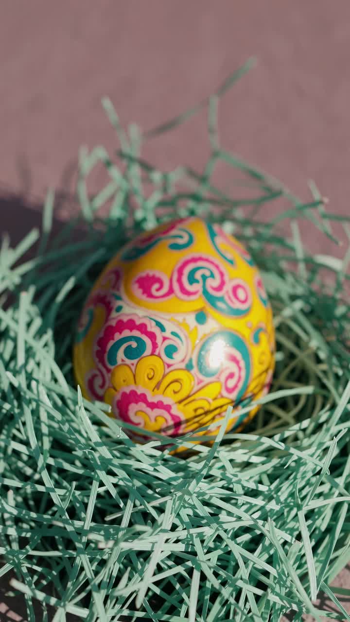 Brightly colored easter egg with yellow, pink, blue and white floral decorations resting on a light blue artificial decorative nest on a pink background