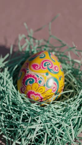 brightly colored easter egg with yellow, pink, blue and white floral decorations resting on a light blue artificial decorative nest on a pink background