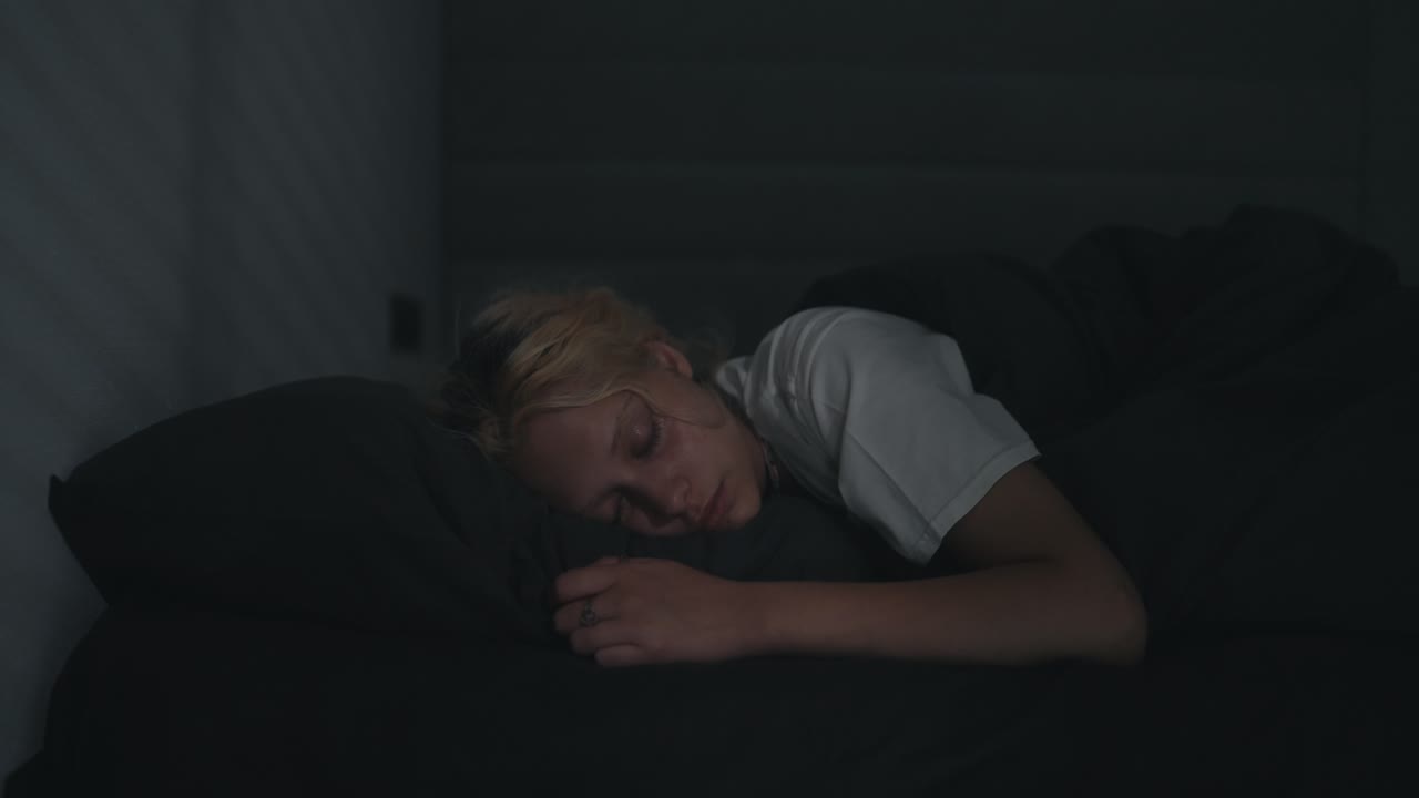 una joven rubia en una camiseta blanca duerme en una cama gris en una habitación oscura