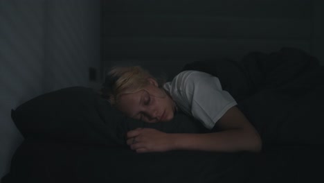 A-young-blonde-girl-in-a-white-T-shirt-sleeps-in-a-gray-bed-in-a-dark-room