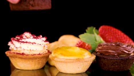 a hand reaches toward and picks up a small pastry from an assortment of colorful desserts, including tarts, cupcakes, brownies, and strawberries, under bright studio lighting with a black background