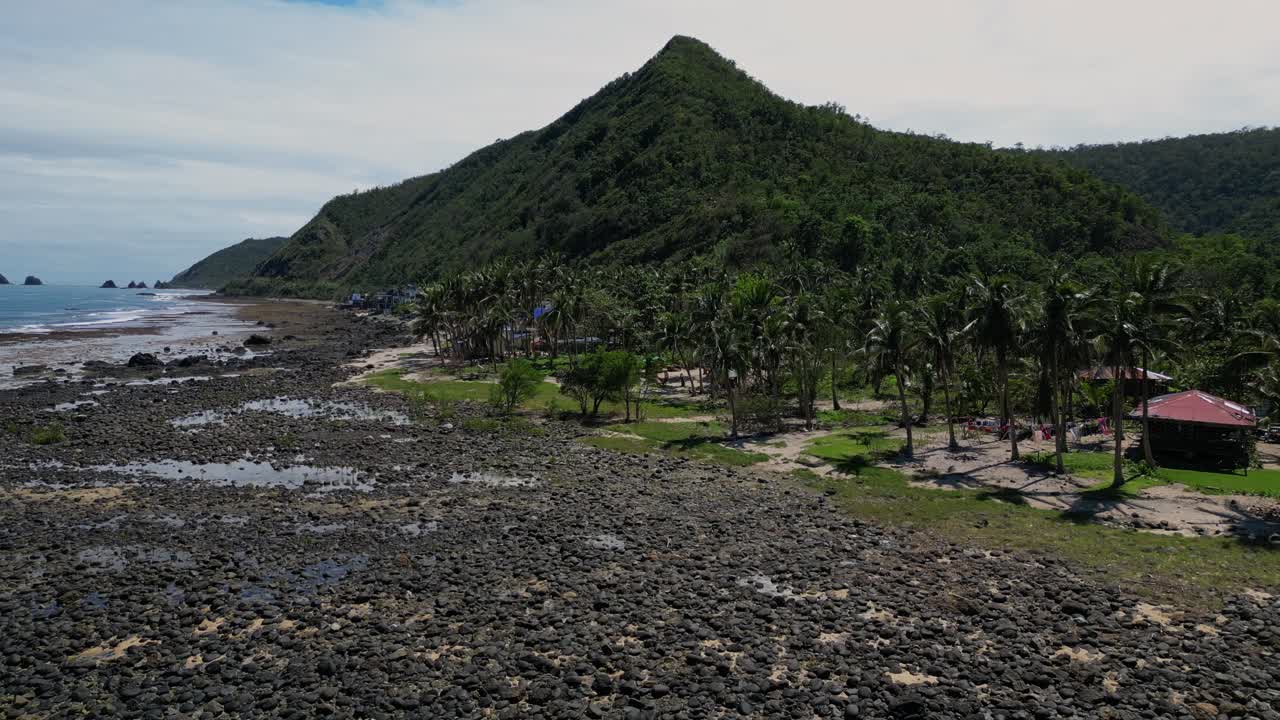 A dolly-out drone shot reveals the shore with palm trees, huts, and houses lining the coastline. A majestic mountain rises above the serene ocean, creating a tranquil, tropical landscape.