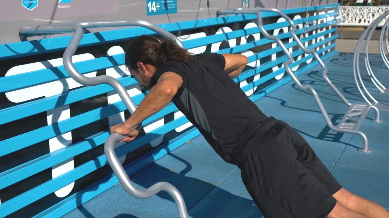 Man with dark hair does ten push ups on bars at outdoor fitness course, medium shot