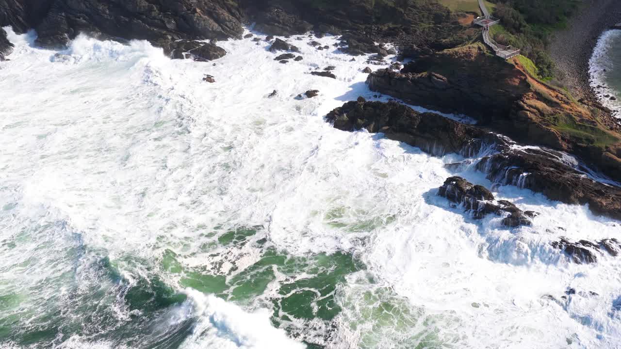 Aerial view of powerful ocean waves hitting rocky cliffs at Byron Bay, Australia. Bright daylight enhances the dynamic coastal scene