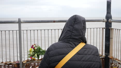 A-pensive-lonely-sad-mourning-person-in-a-hooded-jacket-stands-by-a-seaside-railing,-observing-a-bouquet-of-flowers-placed-in-remembrance-of-loss-and-bereavement-by-the-ocean