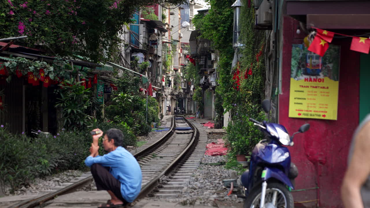 Old Asian man squats on metal tracks at Train Street, with pedestrians walking below tight buildings