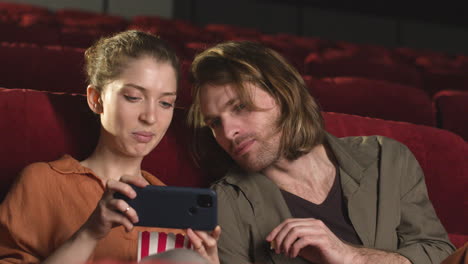 pareja feliz buscando algo interesante en el teléfono móvil mientras se sientan en el cine antes de que comience la película