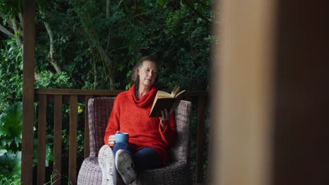mujer caucásica mayor relajándose en el jardín, sentada con los pies en alto, bebiendo café y leyendo un libro