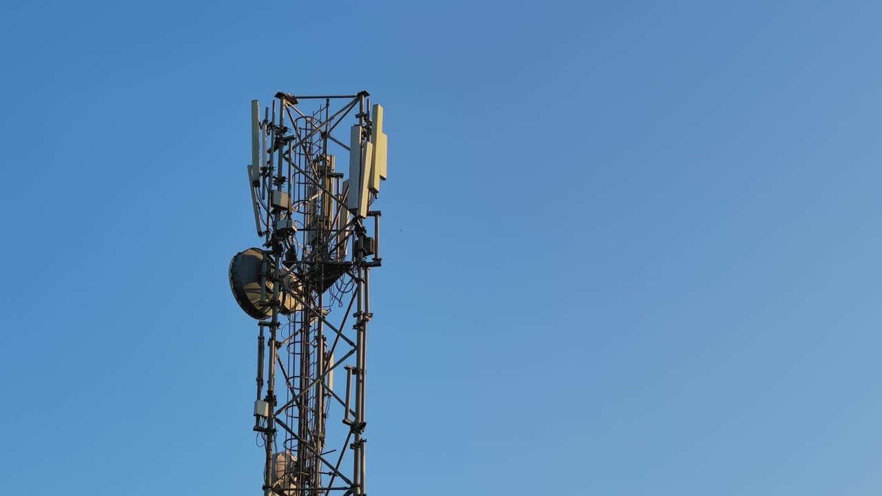 Vista ravvicinata di una torre di comunicazione mobile con antenne a pannello montate, cavi e parabola a microonde contro un cielo azzurro limpido, che rappresenta la moderna infrastruttura di rete wireless.