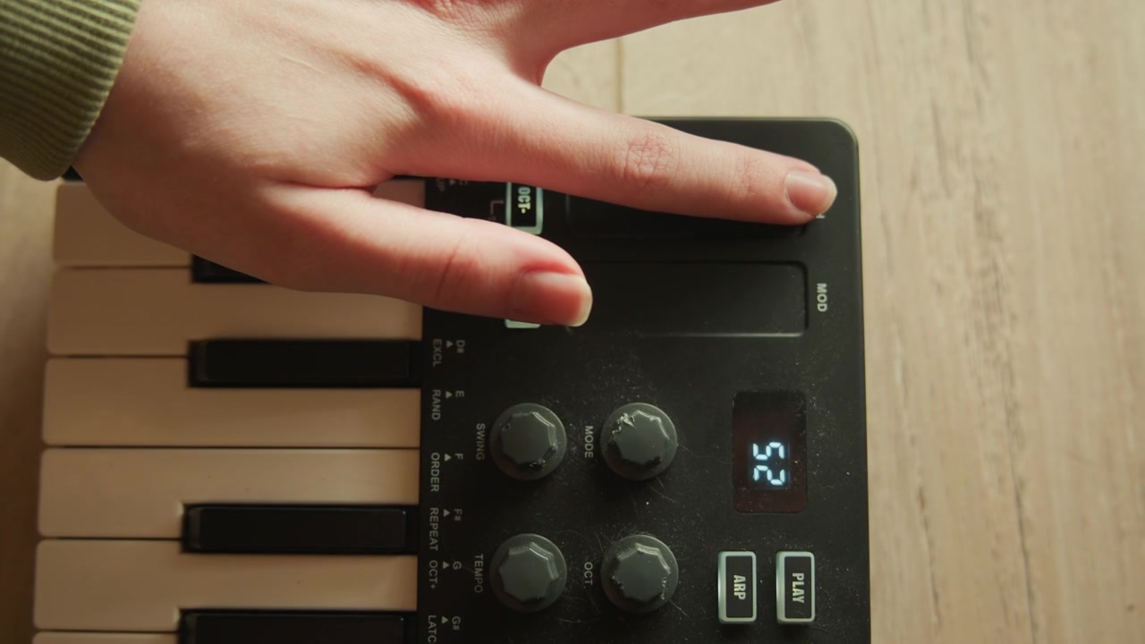Gros plan en plongée montrant la main gauche manipulant un contrôleur de clavier MIDI compact, l'index appuyant sur un bouton rectangulaire tandis que les autres doigts planent au-dessus des touches blanches brillantes dans une lumière de studio chaleureuse