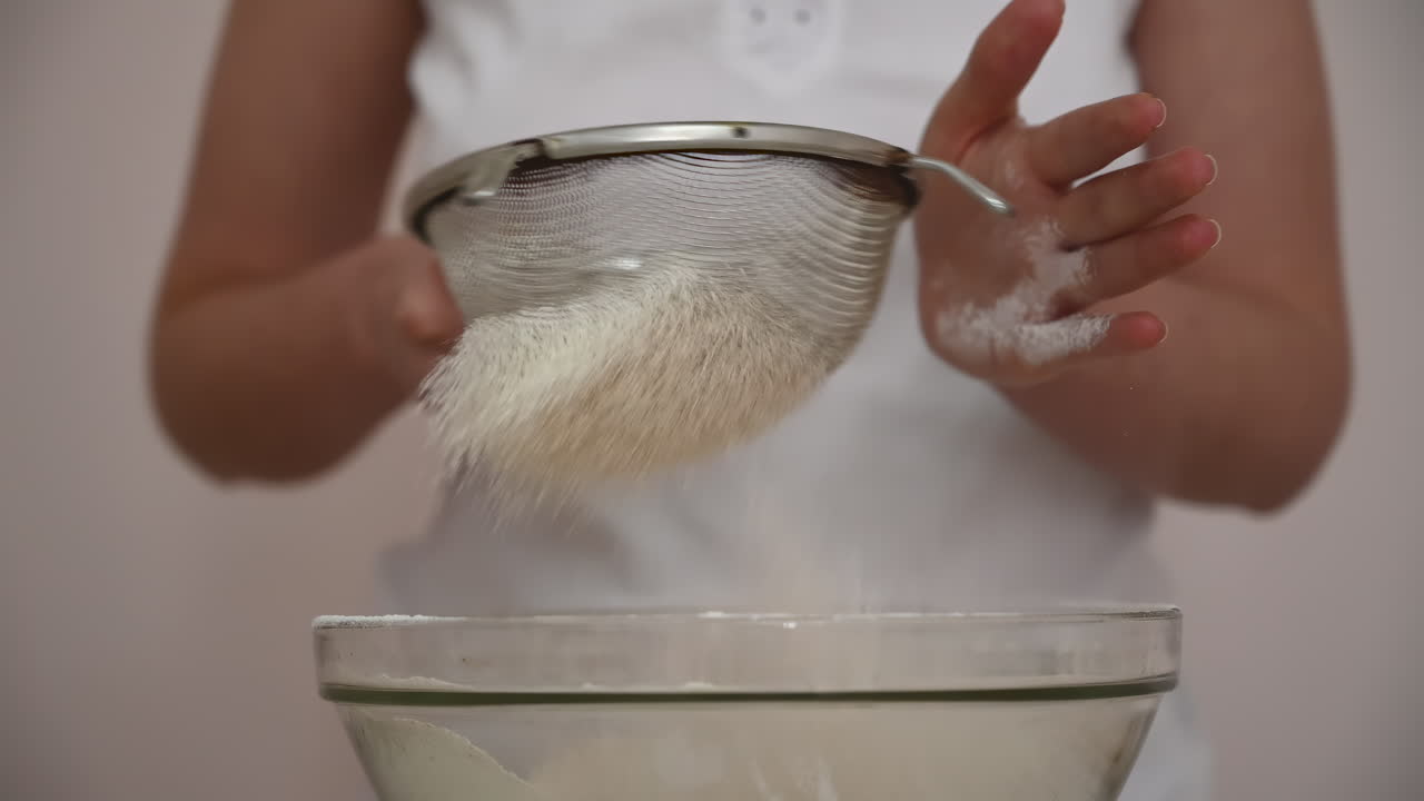 Person sifting flour into a glass bowl while baking in the kitchen