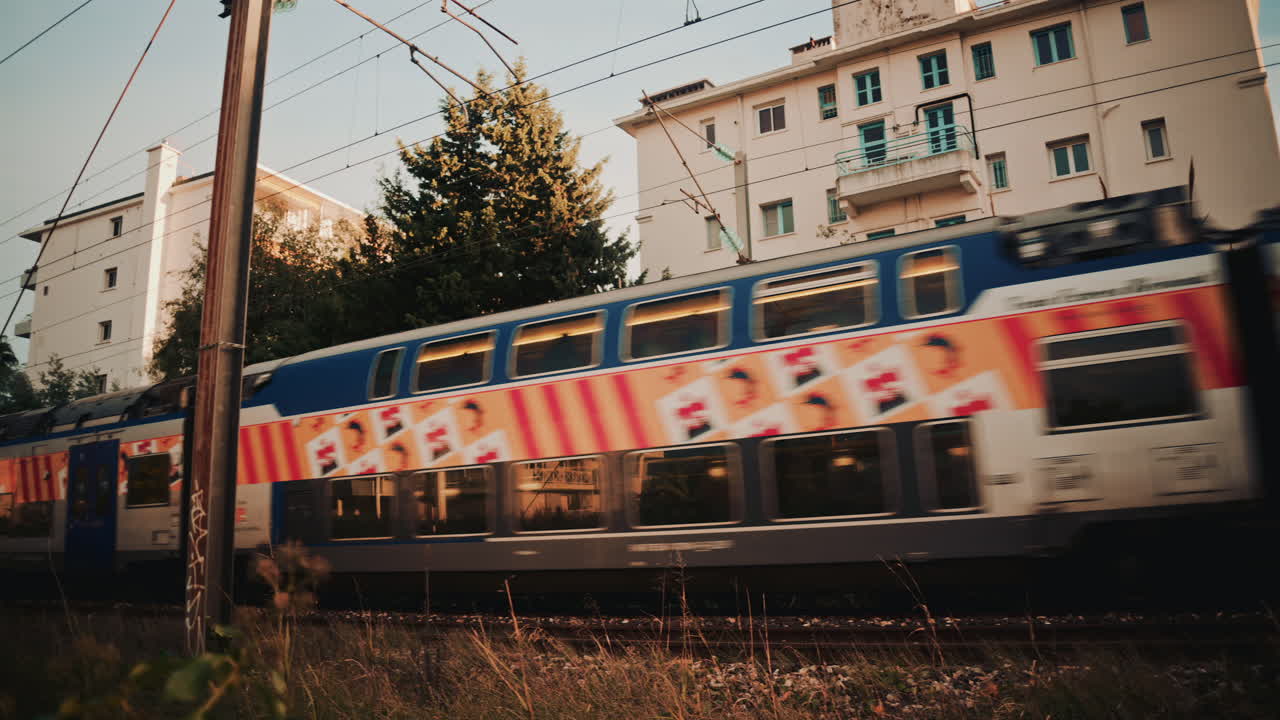 A blue commuter train speeds past a residential building, creating strong motion blur against the still architecture