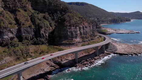 Aerial-view-of-red-cars-driving-on-Sea-Cliff-Bridge,-sunny-day,-grand-pacific-drive,-New-South-Wales,-Australia---forward-drone-shot