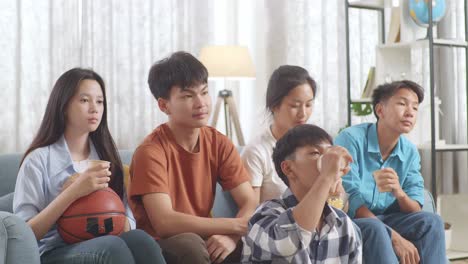 asian teenagers cheering, eating and drink while watching basketball game on tv at home