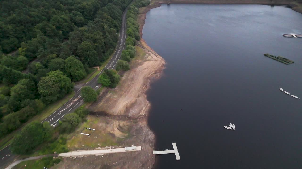 Drone footage glides above Ladybower Reservoir’s shoreline, revealing docks, boats, and a winding road bordered by lush summer greenery under soft daylight