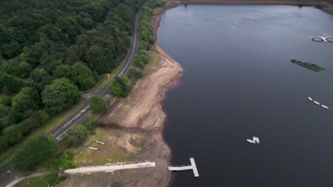drone footage glides above ladybower reservoir’s shoreline, revealing docks, boats, and a winding road bordered by lush summer greenery under soft daylight