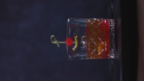 old fashioned cocktail drink chilean version with bourbon whiskey, sugar, ice, bitter dashes, maraschino cherry in dark background and selective focus, vertical video