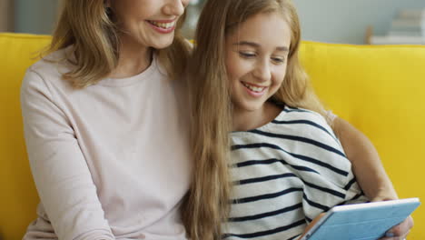 Vista-De-Cerca-De-La-Madre-Y-La-Hija-Rubias-Sonriendo-Y-Viendo-Algo-En-La-Tableta-Mientras-Se-Sientan-En-El-Sofá-En-La-Sala-De-Estar