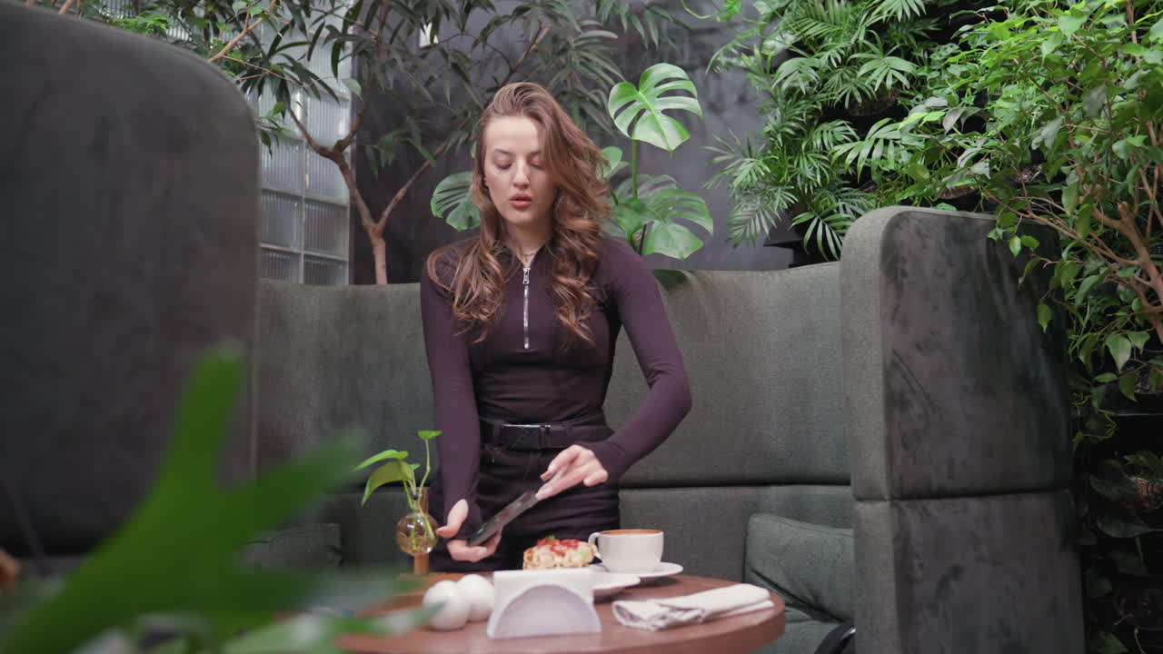 Lady stands in cozy indoor setting, holding phone in landscape mode as she photographs waffles topped with cream and strawberries beside latte with foam art on wooden table surrounded by lush plants