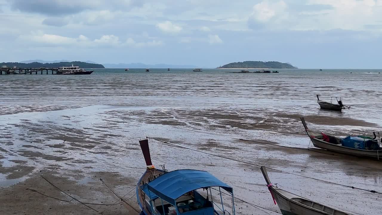 Cropped view of boats resting on a muddy beach during low tide, with distant islands under cloudy skies.