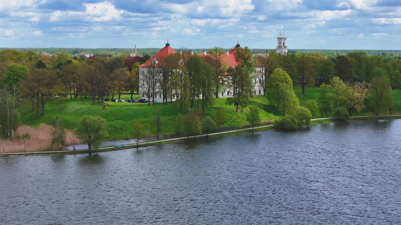 Biržai Castle in late spring: a wide-angle view from across a serene lake, capturing the white-walled, red-roofed historical building amidst vibrant green trees under a partly cloudy sky