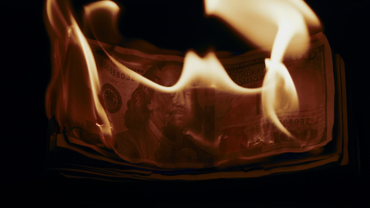 Slow motion medium shot of a stack of prop money as it catches on fire and burns.
