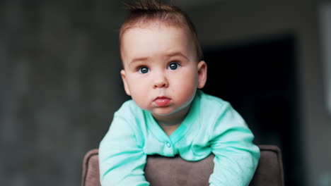 grey-eyed toddler with sticking hair stands on a chair with serious face expression. lovely kid looking around. blurred backdrop.