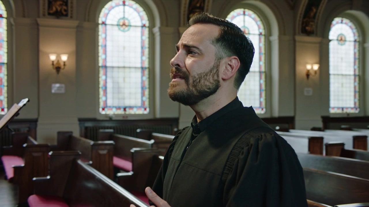 La caméra recule, montrant un homme en robe cléricale noire dirigeant un chant religieux et gesticulant dans une église, les bancs étant visibles.
