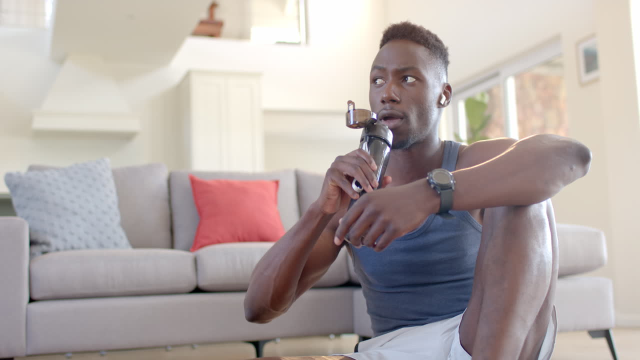 Focused african american man drinking water and using headphones in sunny living room, slow motion